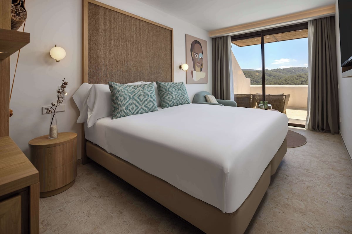 Une chambre d’hôtel avec un grand lit, une petite table de chevet, une lampe fixée au mur, un tableau et un balcon avec vue.
