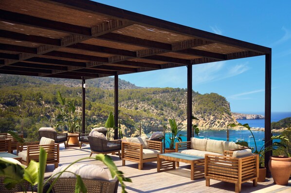 A covered outdoor seating area with wicker furniture and potted plants, overlooking a coastal landscape with hills and the sea. - Cala San Miguel Ibiza Resort, Curio Collection by Hilton (Sant Joan de Labritja)