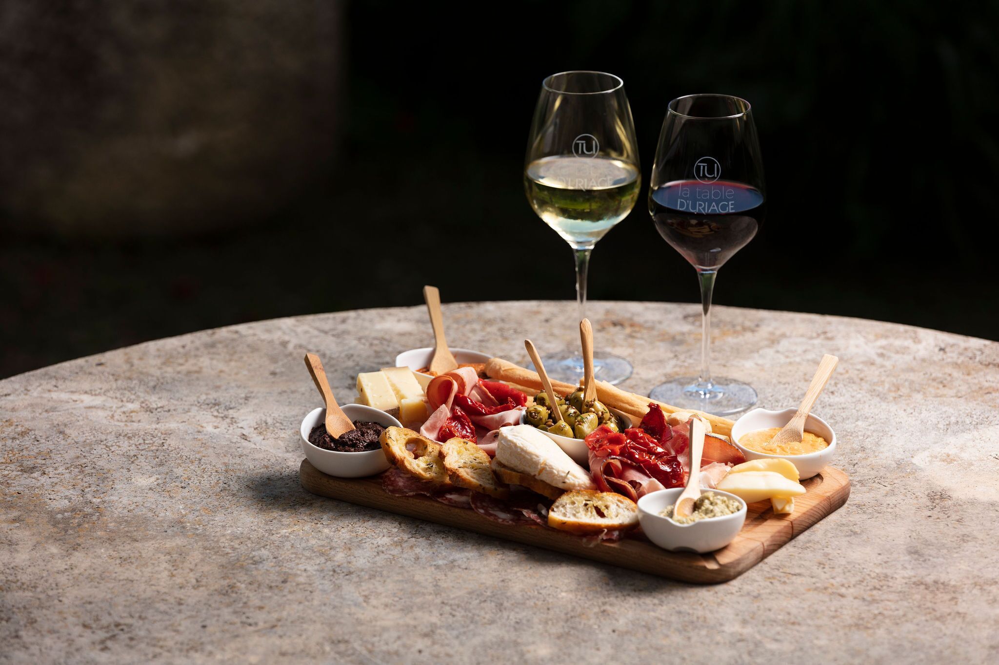 Una tabla de embutidos con varios quesos, carnes y acompañamientos sobre una tabla de madera, junto con dos copas de vino blanco.