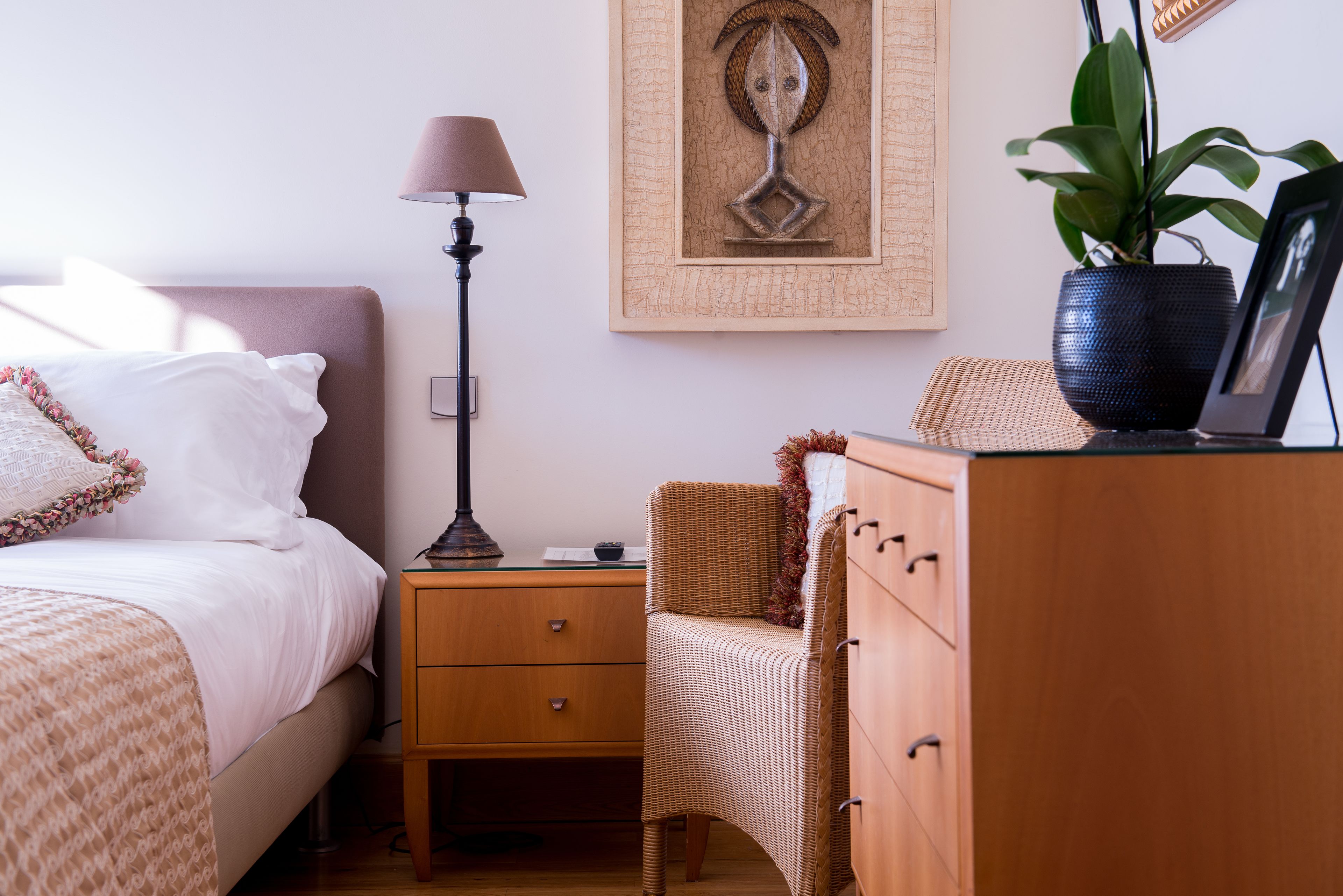 Un dormitorio ordenado con cama, mesita de noche, lámpara, silla, cómoda y una obra de arte enmarcada en la pared.