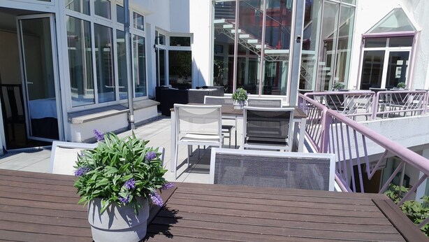 A modern outdoor deck with a wooden floor, white outdoor furniture, and a potted plant with purple flowers. - Hotel Aichtaler Hof (Aichtal)
