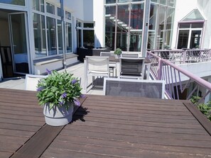 Una terraza moderna al aire libre con piso de madera, mobiliario exterior blanco y una maceta con flores moradas.