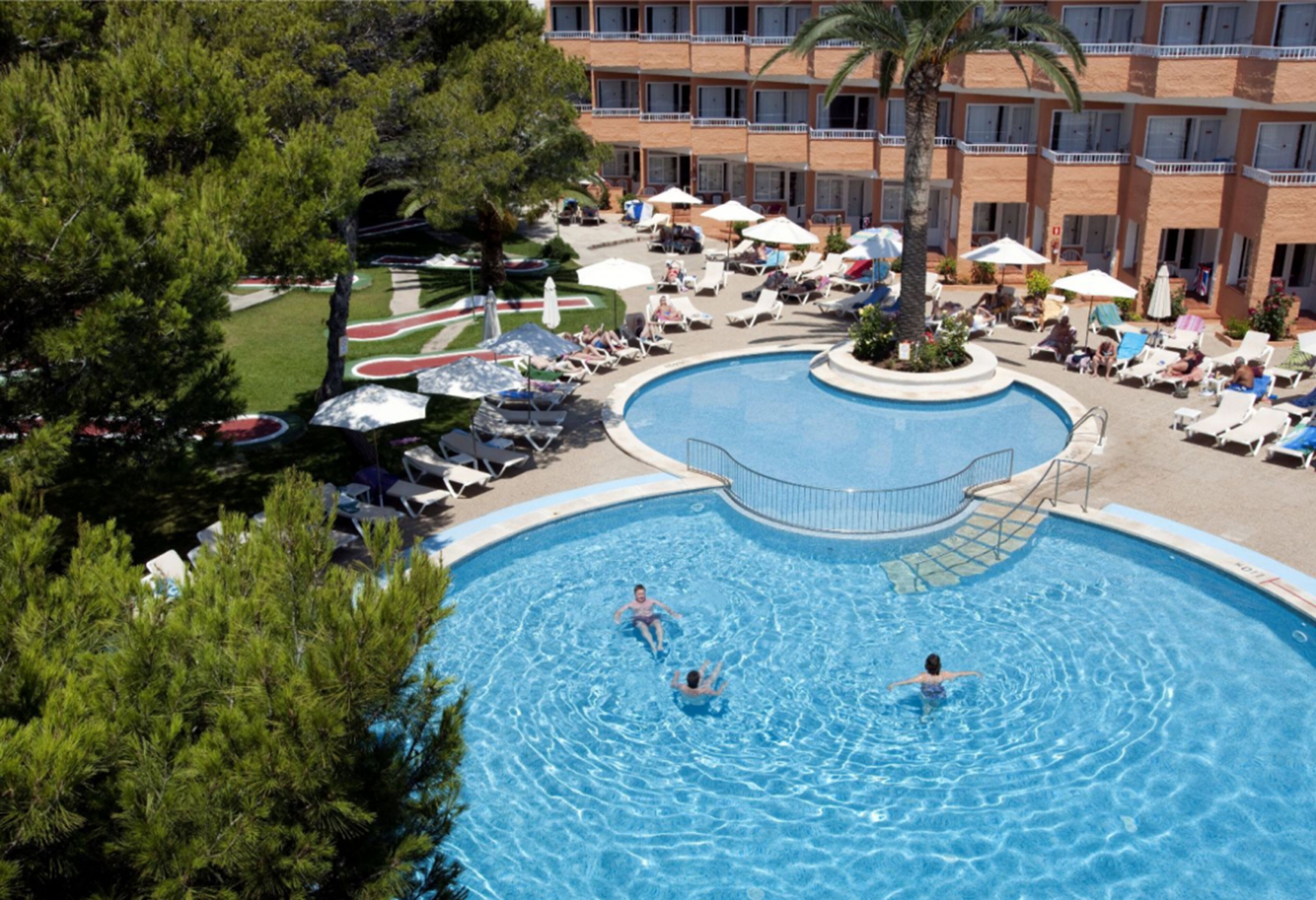 A swimming pool with lounge chairs and umbrellas, surrounded by a landscaped area and a multi-story building.