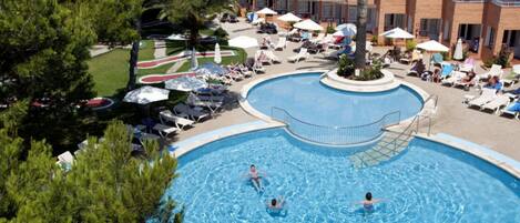 A swimming pool with lounge chairs and umbrellas, surrounded by a landscaped area and a multi-story building.