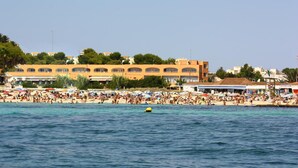 Una playa concurrida con gente y sombrillas, un mar azul y despejado, y edificios al fondo.