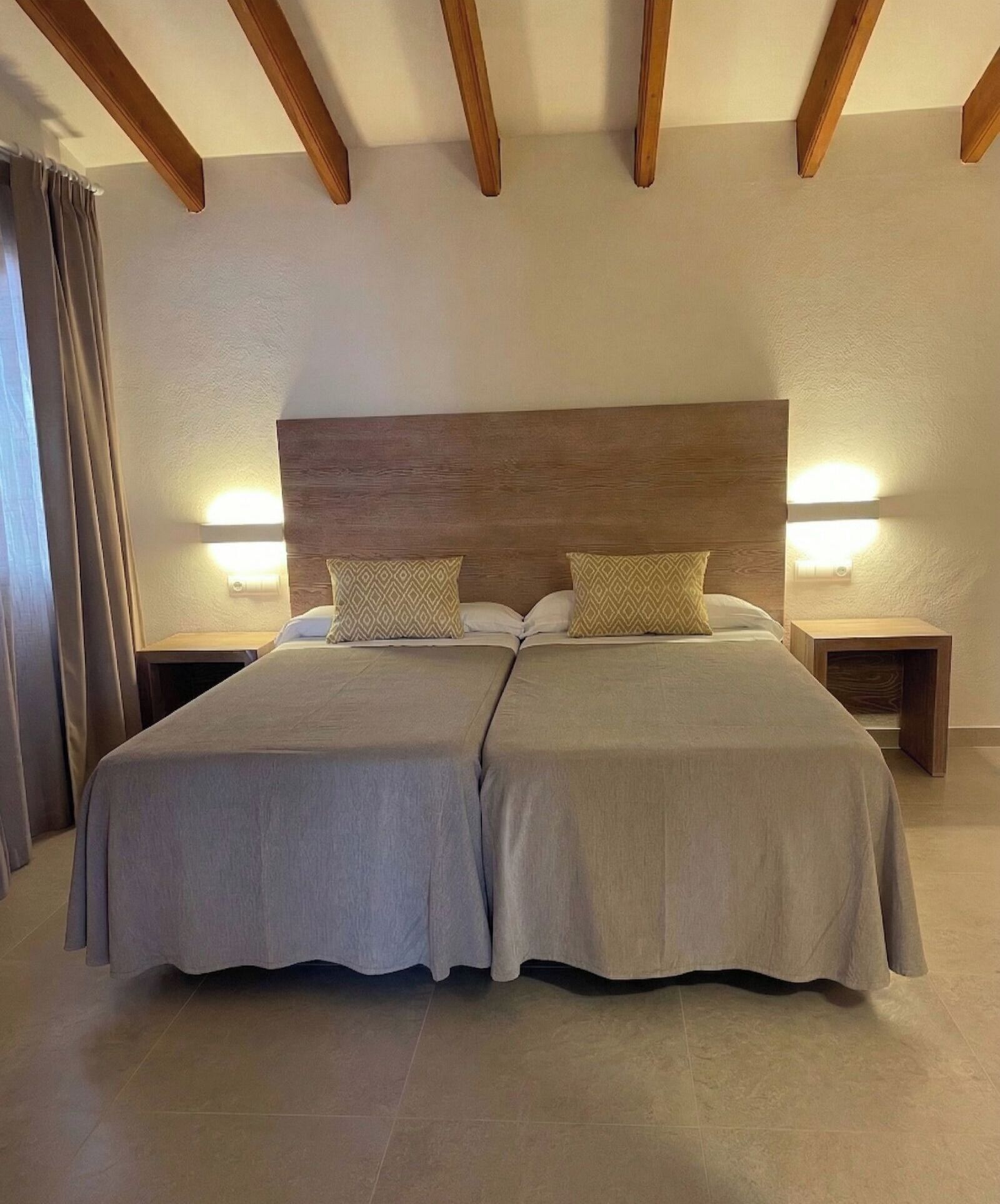 Una cama doble con cabecera de madera, dos mesitas de noche con lámparas y una cortina a la izquierda.