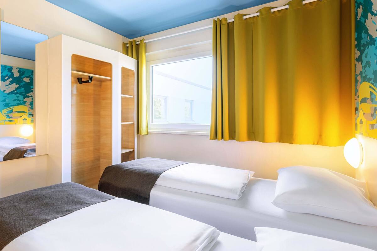Habitación de hotel con dos camas, un armario y una ventana con cortinas.