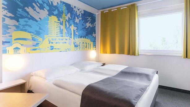 A hotel room with a bed, a desk, a window with curtains, and a wall mural. - B&B Hotel Stuttgart-City (Stuttgart)