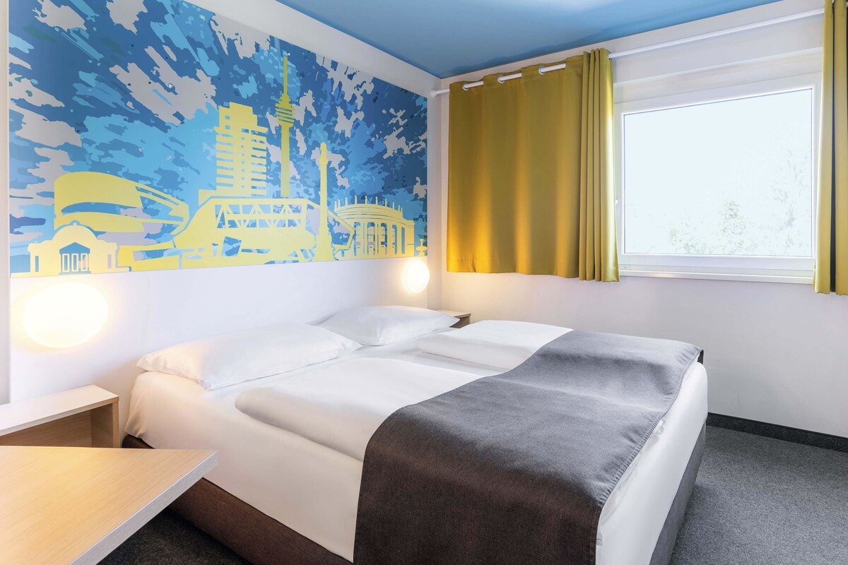 A hotel room with a bed, a desk, a window with curtains, and a wall mural.