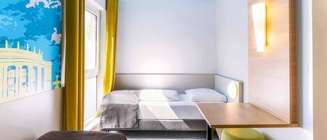 A compact hotel room with a bed, a small desk, a chair, and a wall-mounted light fixture.