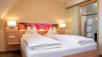A hotel room with a double bed, two bedside lamps, a wooden headboard, a large floral artwork above the bed, and a bathroom with a towel rack and shower.