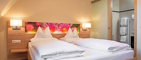 Ein Hotelzimmer mit Doppelbett, zwei Nachttischlampen, einem Holz-Kopfteil, einem großen Blumenbild über dem Bett und einem Badezimmer mit Handtuchhalter und Dusche.