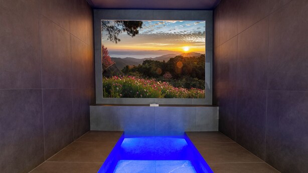 Une salle de spa dotée d’une grande fenêtre offrant une vue sur le coucher du soleil, d’un sol éclairé de lumière bleue et de murs recouverts de carreaux.