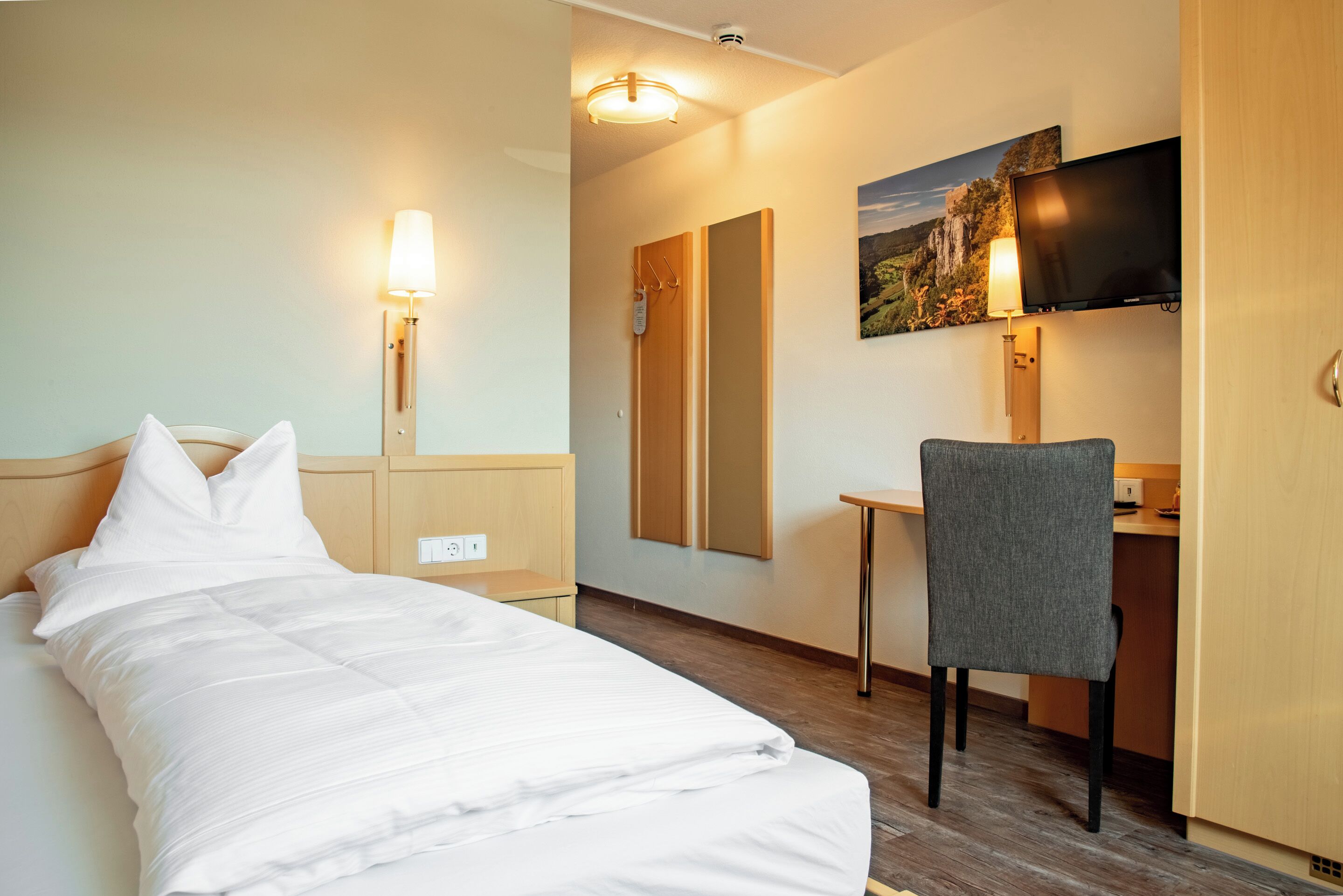 A hotel room with a bed, desk, chair, and a TV mounted on the wall.