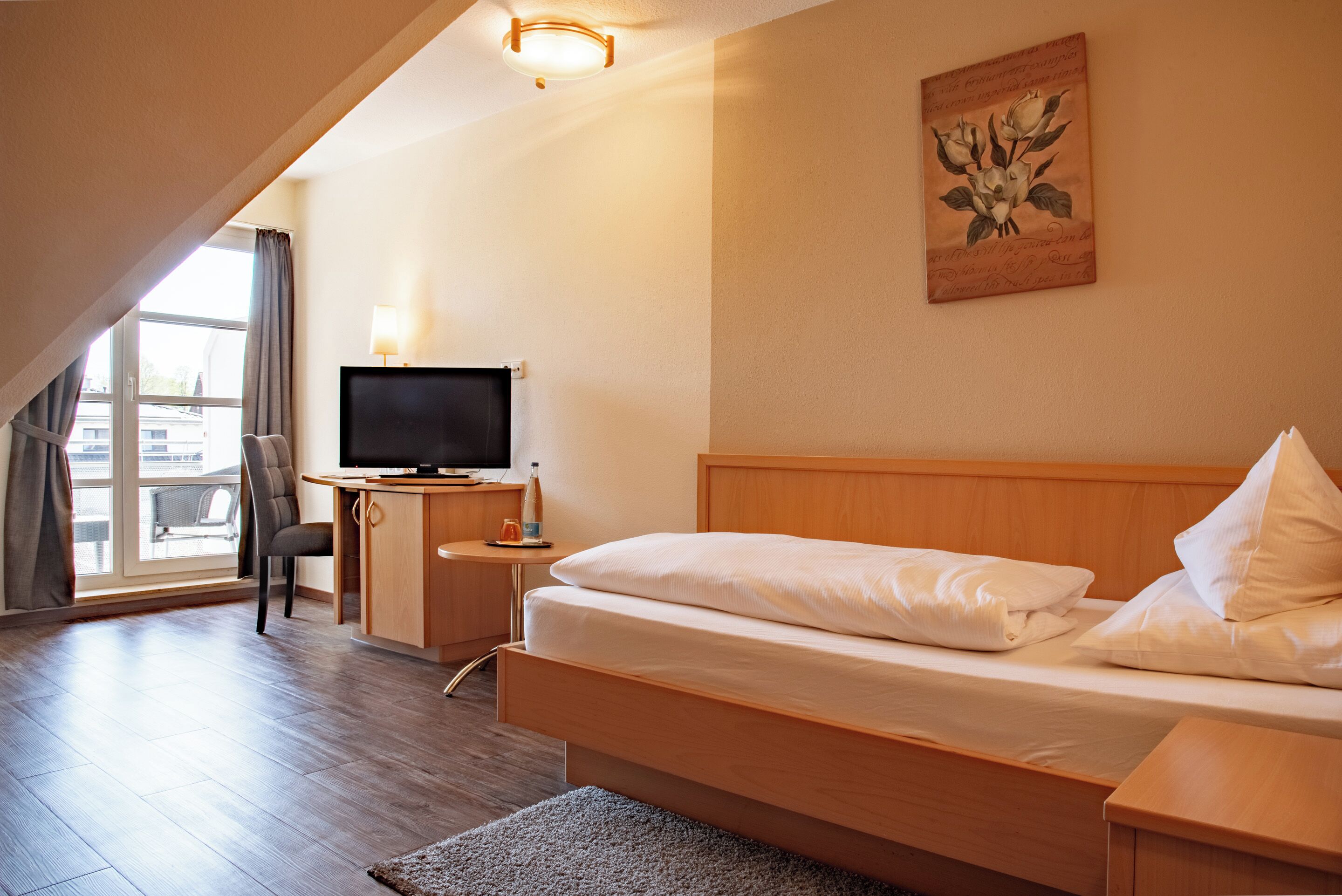 A hotel room with a bed, a desk with a TV, a chair, and a window with curtains.