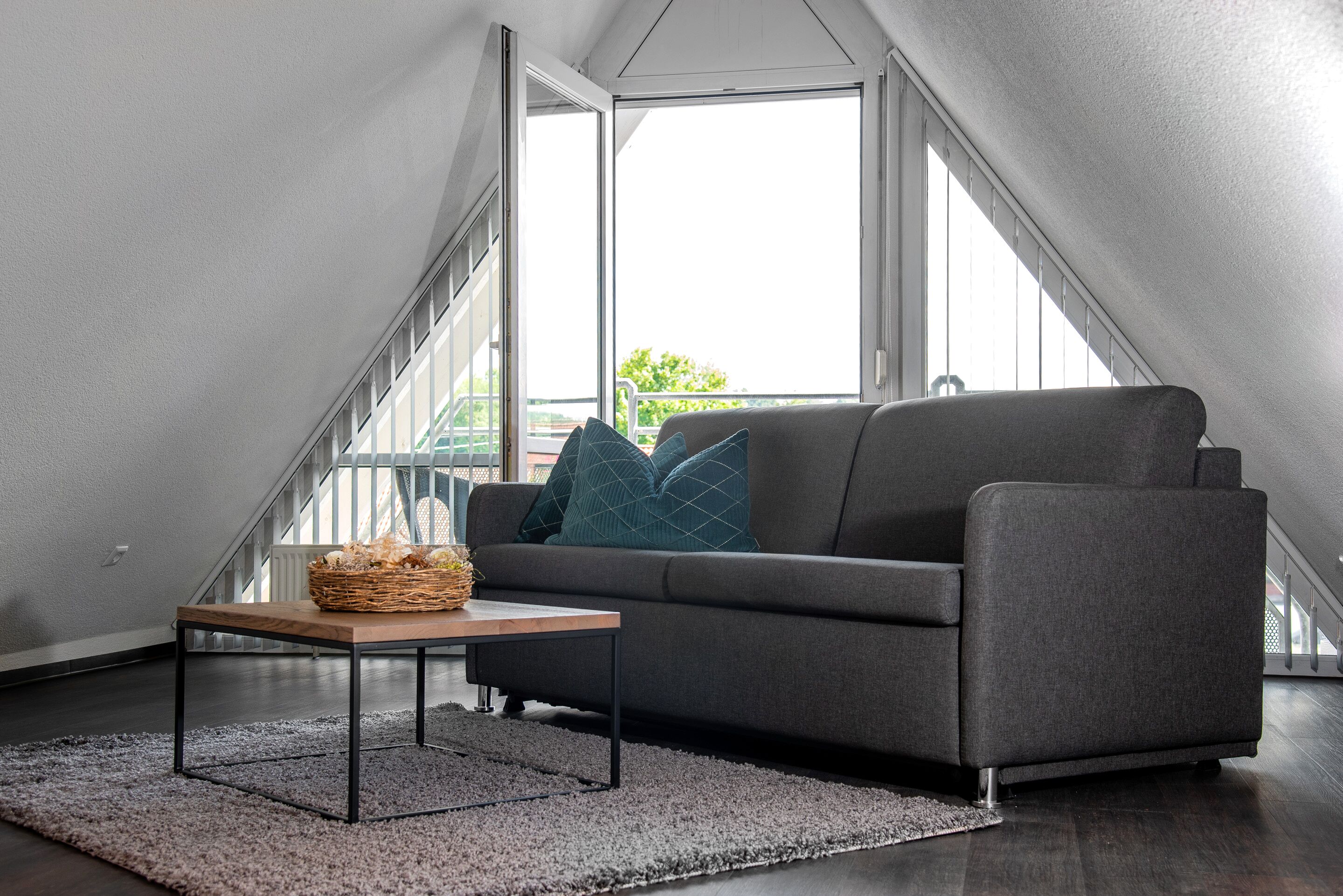 A modern living room with a grey sofa, a wooden coffee table, and a grey rug. A basket is on the table. The room has large windows and a view of a balcony.