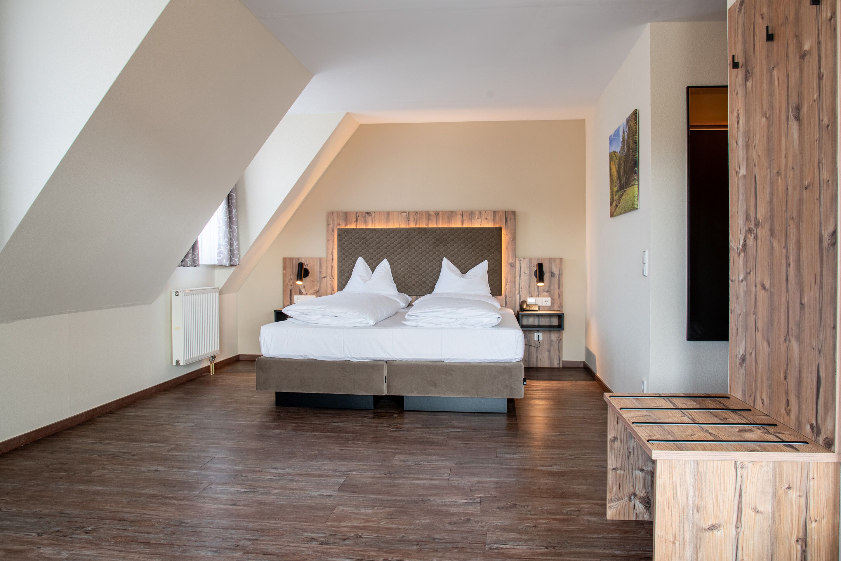 Un dormitorio moderno con techo inclinado, una cama grande con ropa de cama blanca, cabecera de madera y un banco de madera.