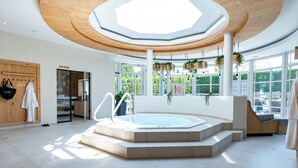 Un espace piscine intérieur moderne, doté d’un grand jacuzzi central, d’un plafond en bois et de grandes fenêtres offrant une vue sur la verdure.