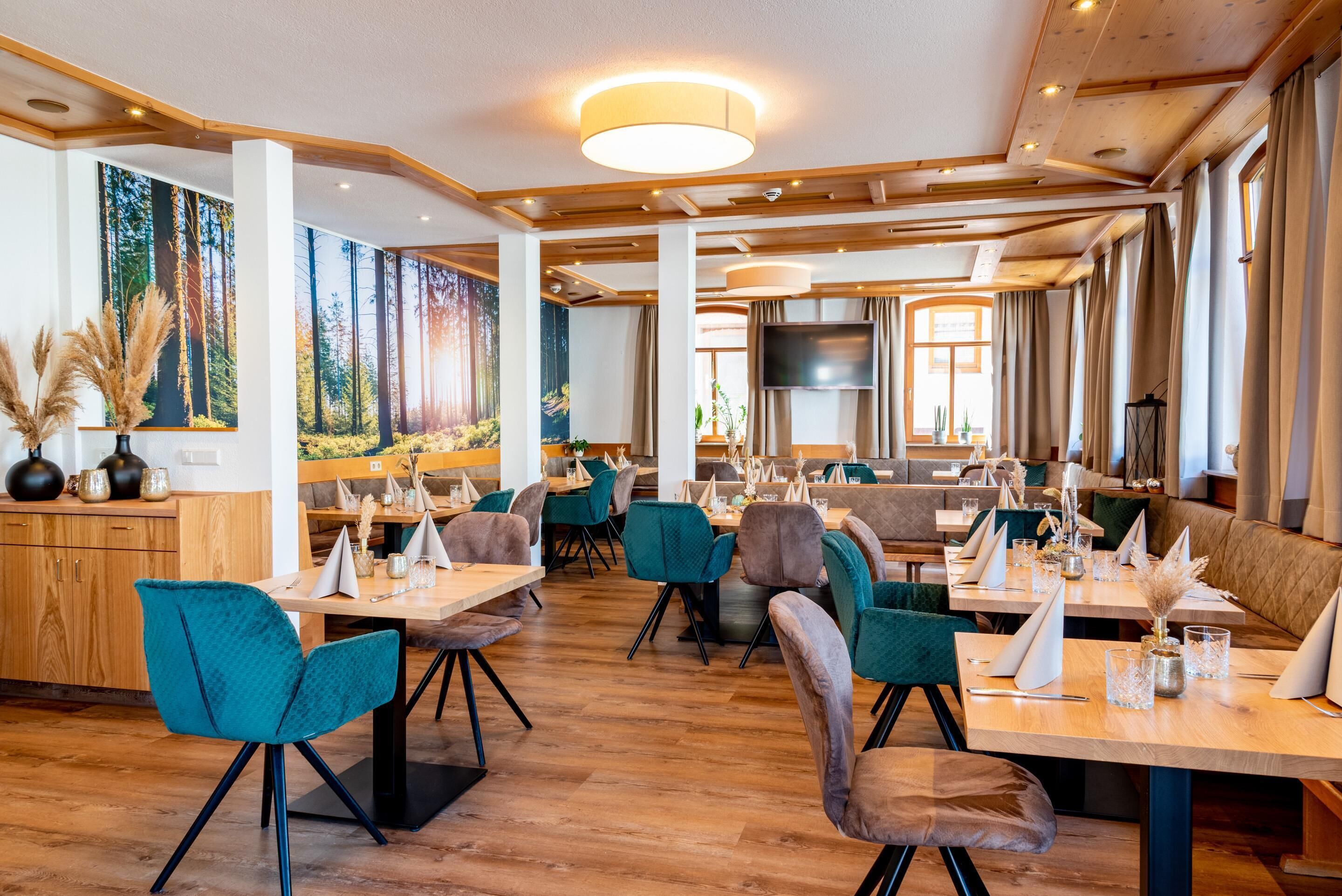 A spacious restaurant with wooden floors, teal chairs, and a large mural of a forest. Tables are set with white napkins and glassware.