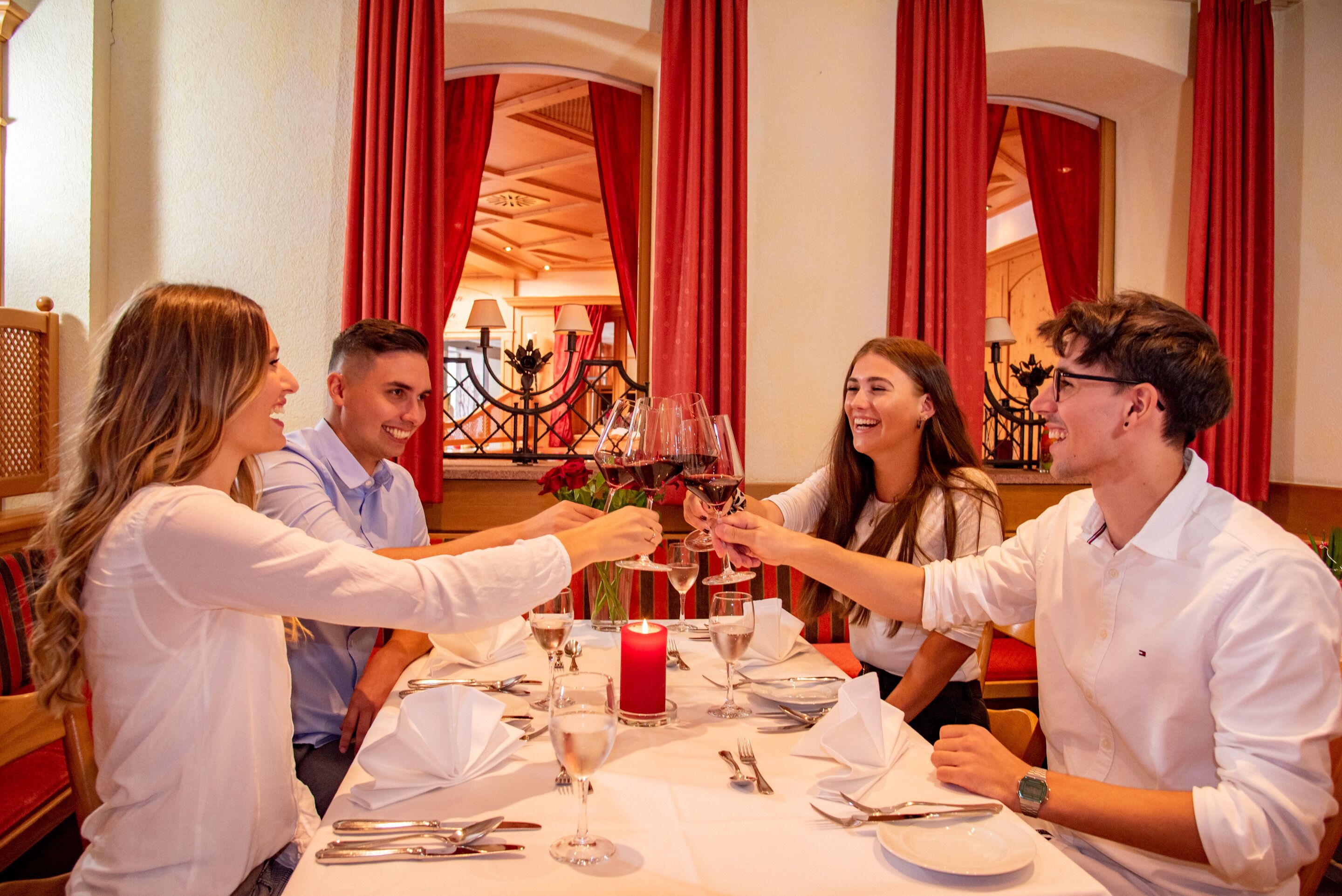 Un grupo de personas sentadas en una mesa con copas de vino y una vela, en una habitación con cortinas rojas y un candelabro.