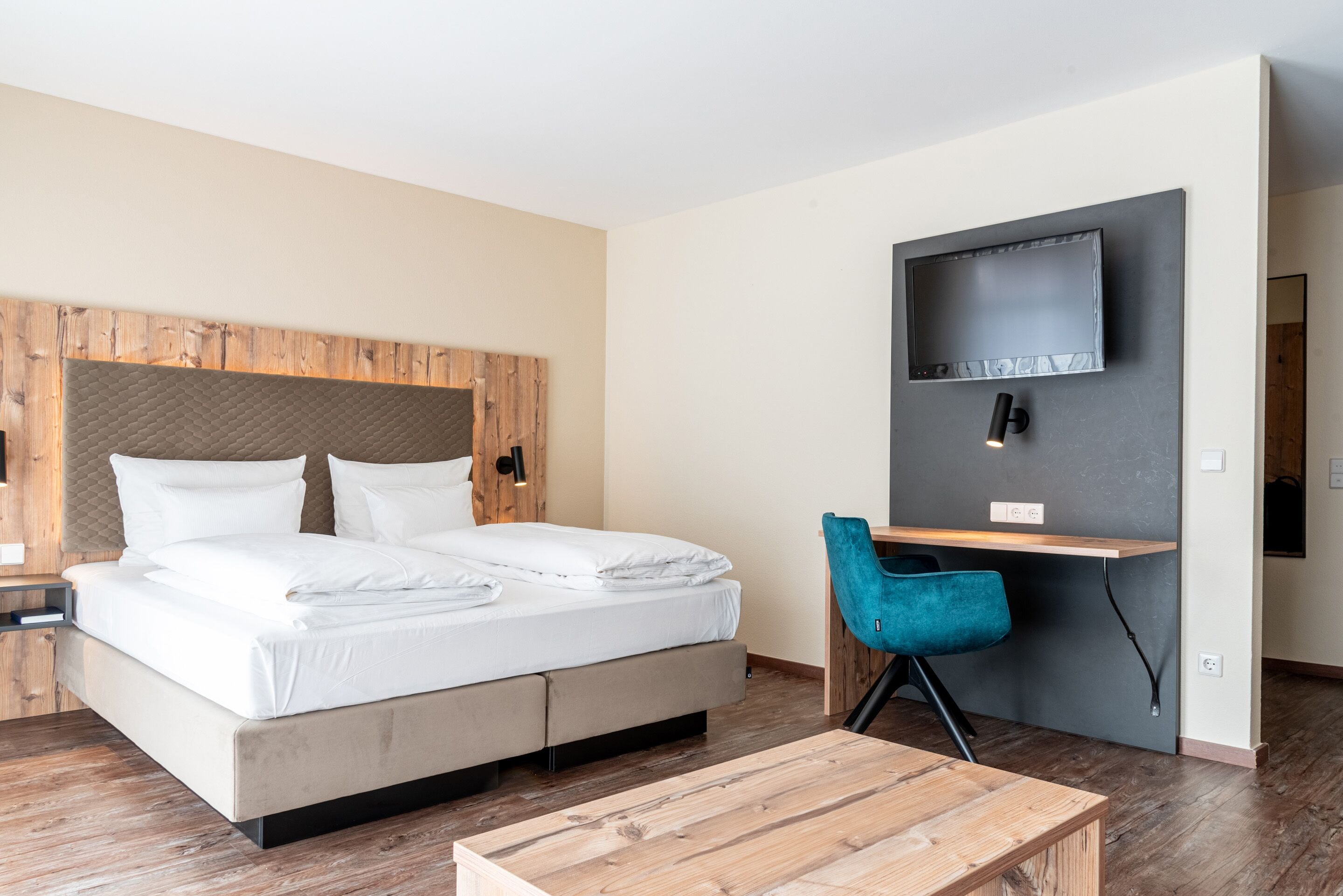 A modern hotel room with a large bed, a desk with a chair, a flat-screen TV mounted on the wall, and a wooden coffee table.