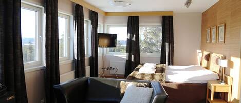 Habitación de hotel con cama, sofá, televisión, una mesa con fruta y ventanas amplias con cortinas.