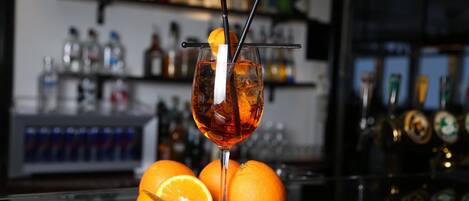 Un vaso de cóctel con pajita y una rodaja de naranja en el borde, sobre una barra con naranjas y una grifo de cerveza al fondo.