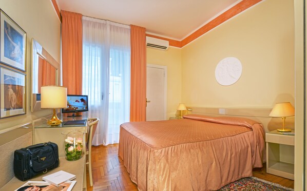 A hotel room with a bed, a desk, a chair, a lamp, a TV, a window with curtains, and a wall clock. - Hotel Aurora Terme (Abano Terme)