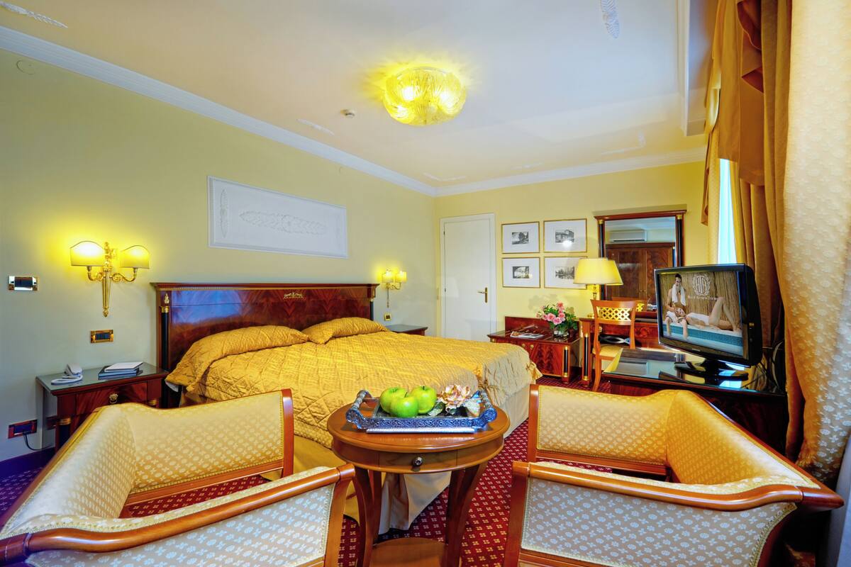 Quarto de hotel com uma cama grande, duas cadeiras, uma mesinha com maçãs, televisão e luminárias fixadas na parede.