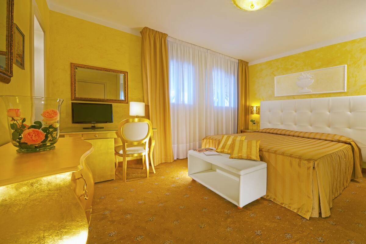 Quarto de hotel com uma cama grande, uma escrivaninha com computador, uma cadeira e um vaso com flores.