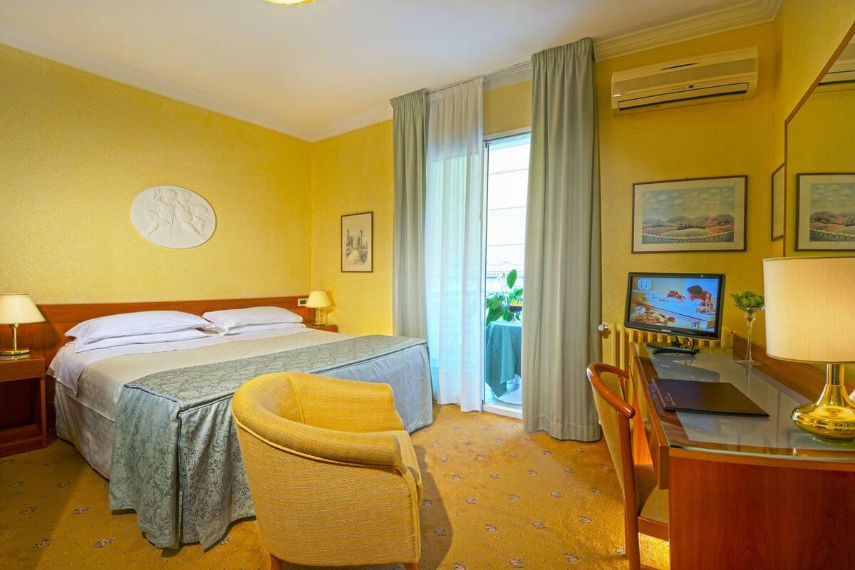 Quarto de hotel com cama, escrivaninha com computador, cadeira, televisão e janela com cortinas.