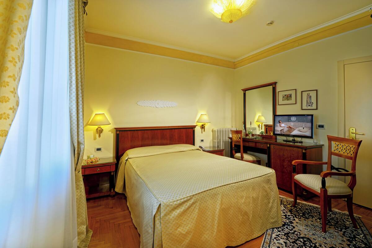 Quarto de hotel com cama, abajures, televisão, escrivaninha e cadeira, e espelho.