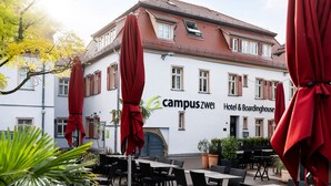 A building with a sign reading "campuszwei Hotel & Boardinghouse" with outdoor seating and red umbrellas. - campuszwei Hotel & Boardinghouse (Ludwigsburg)