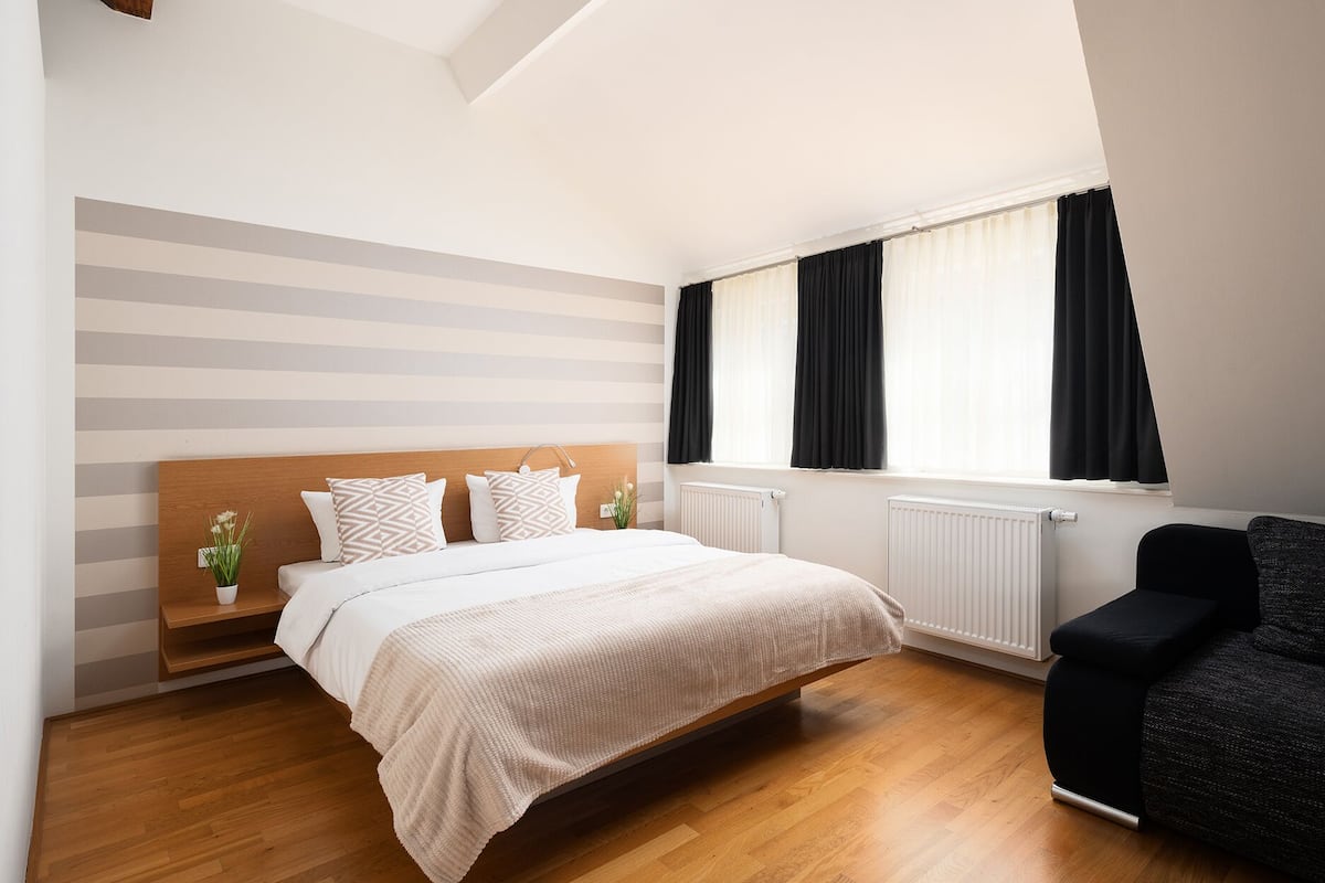 A bedroom with a bed, a wooden headboard, a black chair, a radiator, and a window with curtains.
