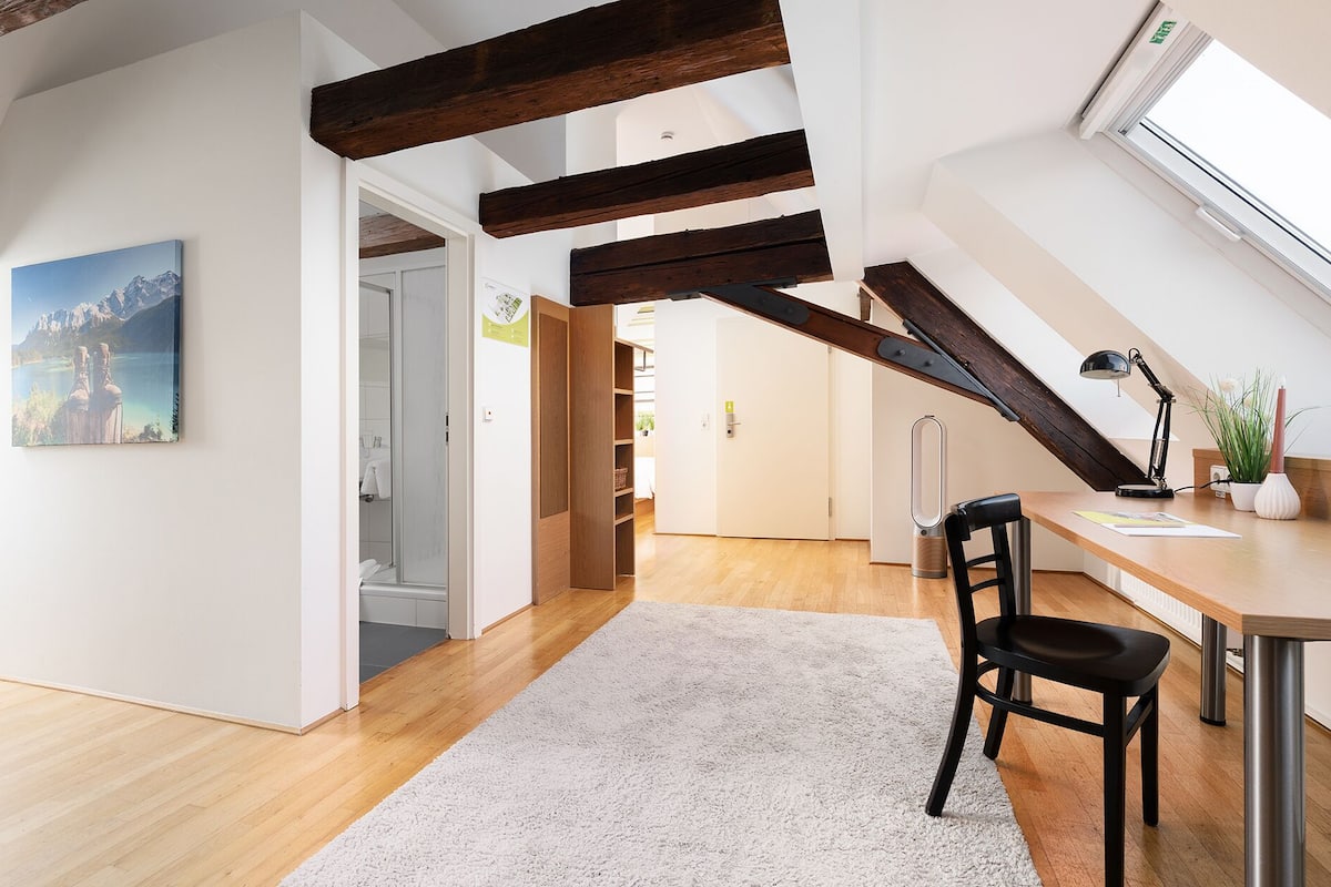 A spacious loft with exposed wooden beams, a wooden desk, a chair, a white rug, and a painting of mountains.