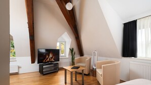 A modern loft bedroom with a sloped ceiling, wooden beams, a television, a sofa, a chair, and a window with curtains. - campuszwei Hotel & Boardinghouse (Ludwigsburg)
