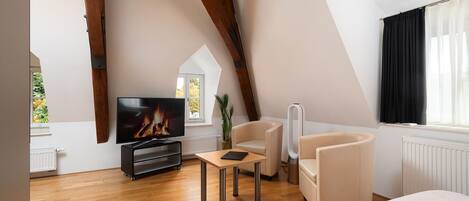 A modern loft bedroom with a sloped ceiling, wooden beams, a television, a sofa, a chair, and a window with curtains.