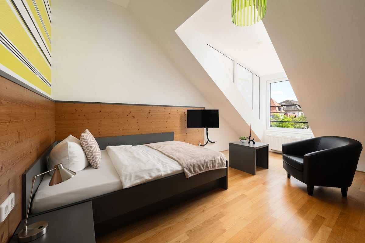 A modern bedroom with a sloped ceiling, a bed, a desk, a chair, and a view of the outside.
