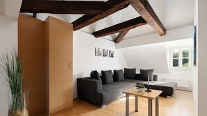 A modern living room with a grey sofa, wooden floor, exposed wooden beams, and a window with a view of greenery. - campuszwei Hotel & Boardinghouse (Ludwigsburg)