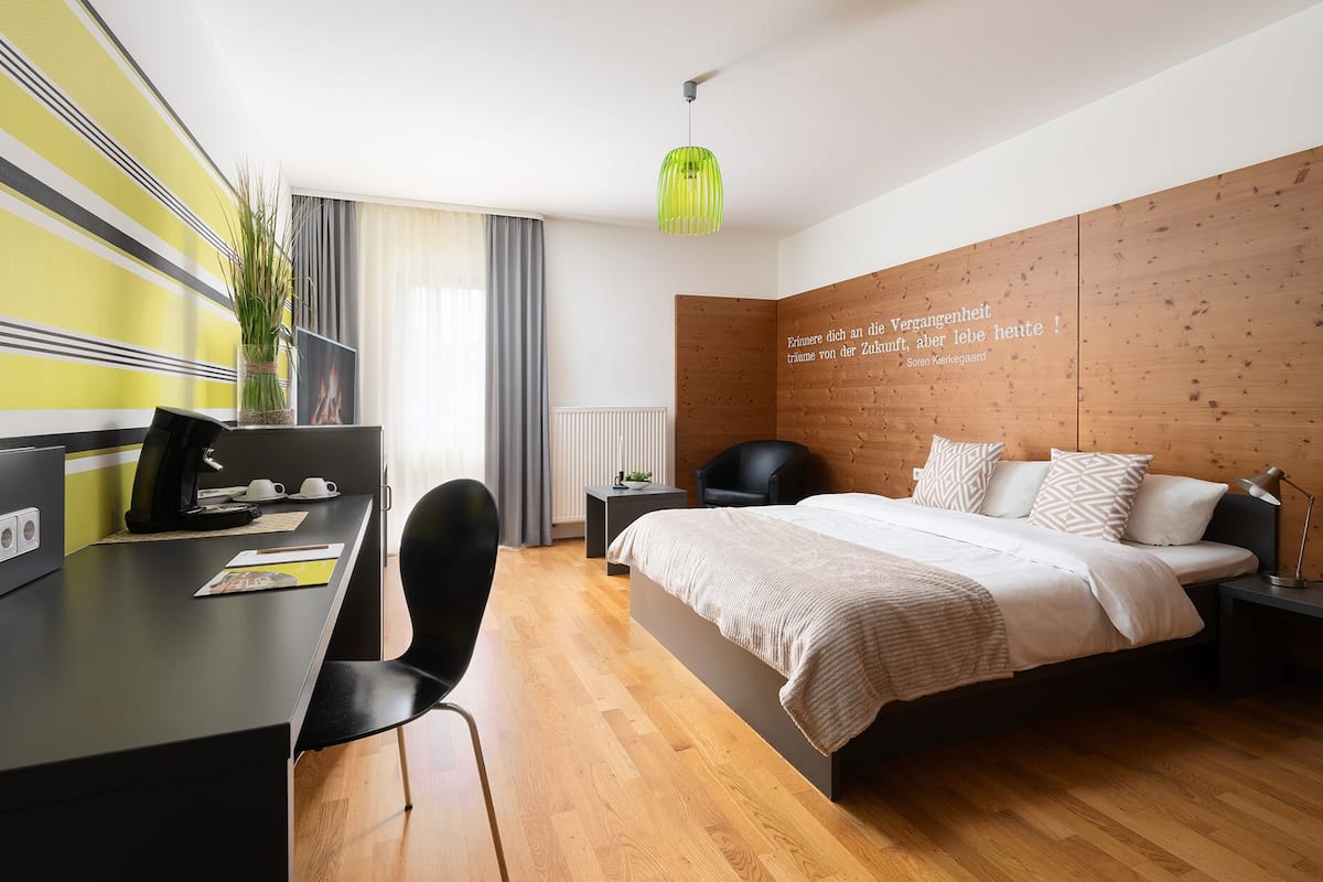 A modern hotel room with a wooden headboard, a bed with a beige blanket, a desk with a laptop, a chair, and a desk lamp.