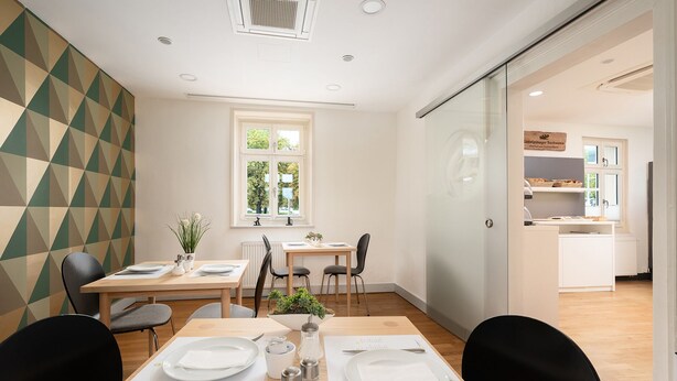 A modern dining area with a geometric-patterned wall, a window with a view, and a kitchen visible through a glass partition. - campuszwei Hotel & Boardinghouse (Ludwigsburg)