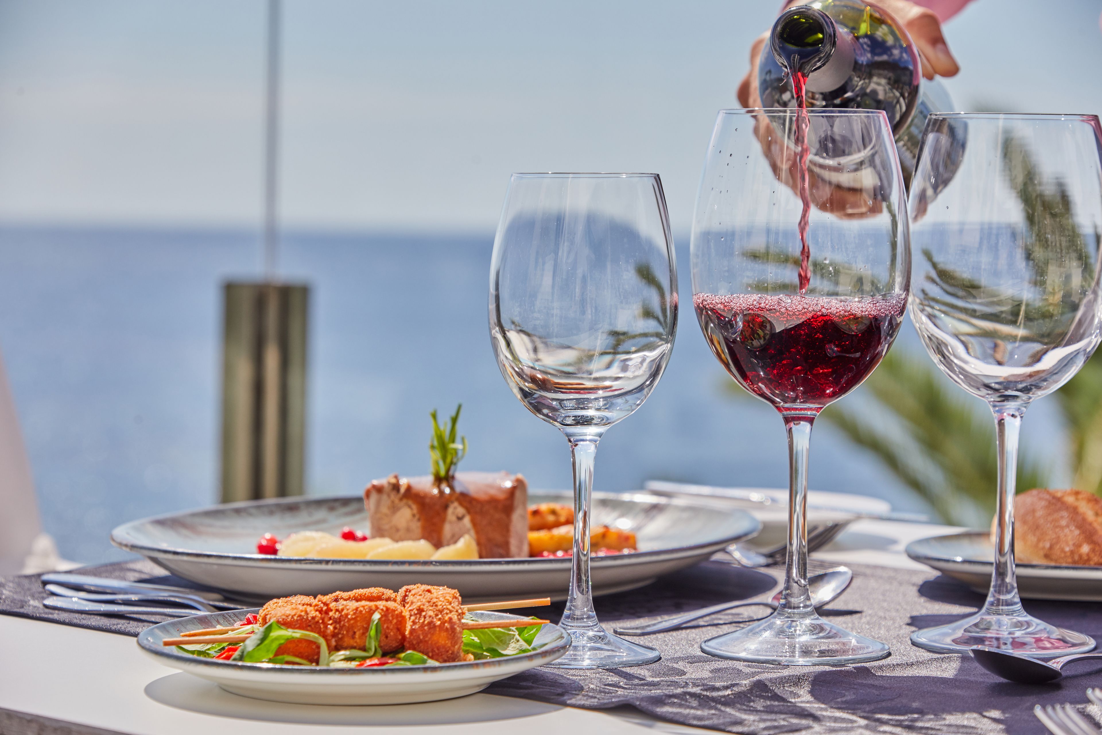 A table with wine glasses, plates of food, and a bottle of red wine being poured.