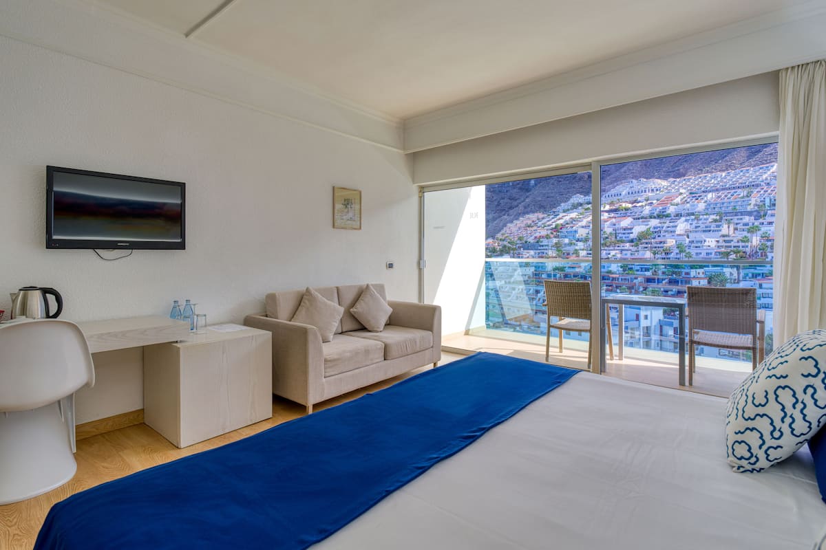 Ein Hotelzimmer mit Bett, Sofa, Schreibtisch, Fernseher und einem Balkon mit Stadtblick.