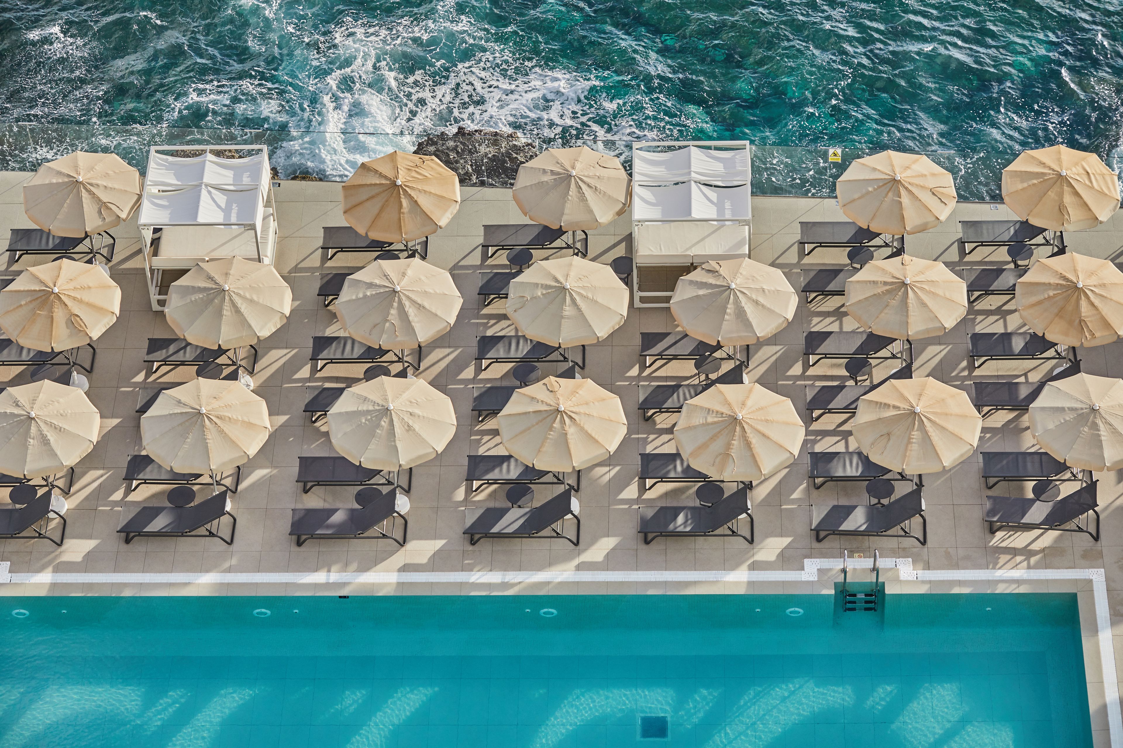 An aerial view of a poolside area with multiple sun loungers and umbrellas, overlooking a body of water.