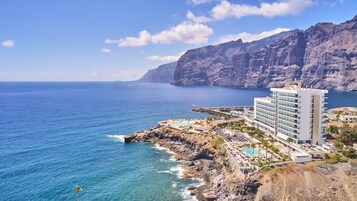 A coastal hotel with a pool, a view of the sea, and a mountainous backdrop.