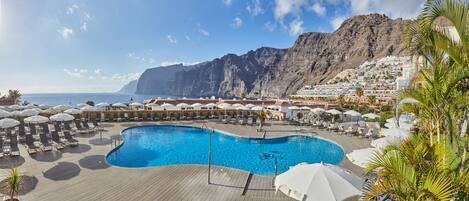 Zwembad van een resort met ligbedden en parasols, omgeven door gebouwen en een berg op de achtergrond.