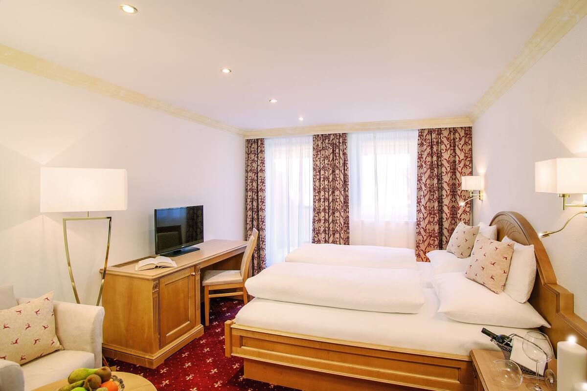 Ein Hotelzimmer mit einem Bett, einem Schreibtisch mit Fernseher, einem Sessel und einem kleinen Tisch mit Flasche und Gläsern.