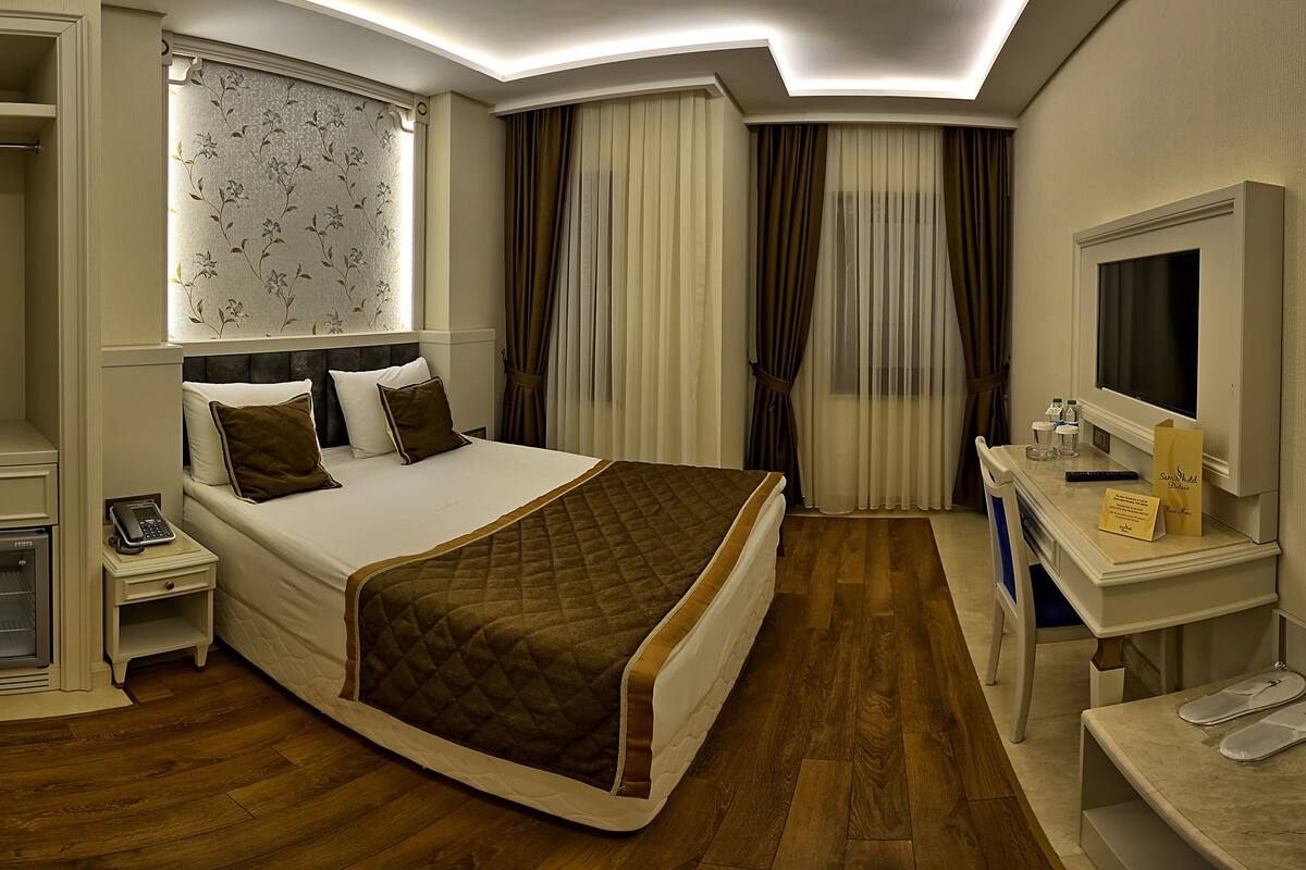 A hotel room with a bed, bedside tables, a desk with a mirror, a TV, and a bathroom with a shower and toilet.