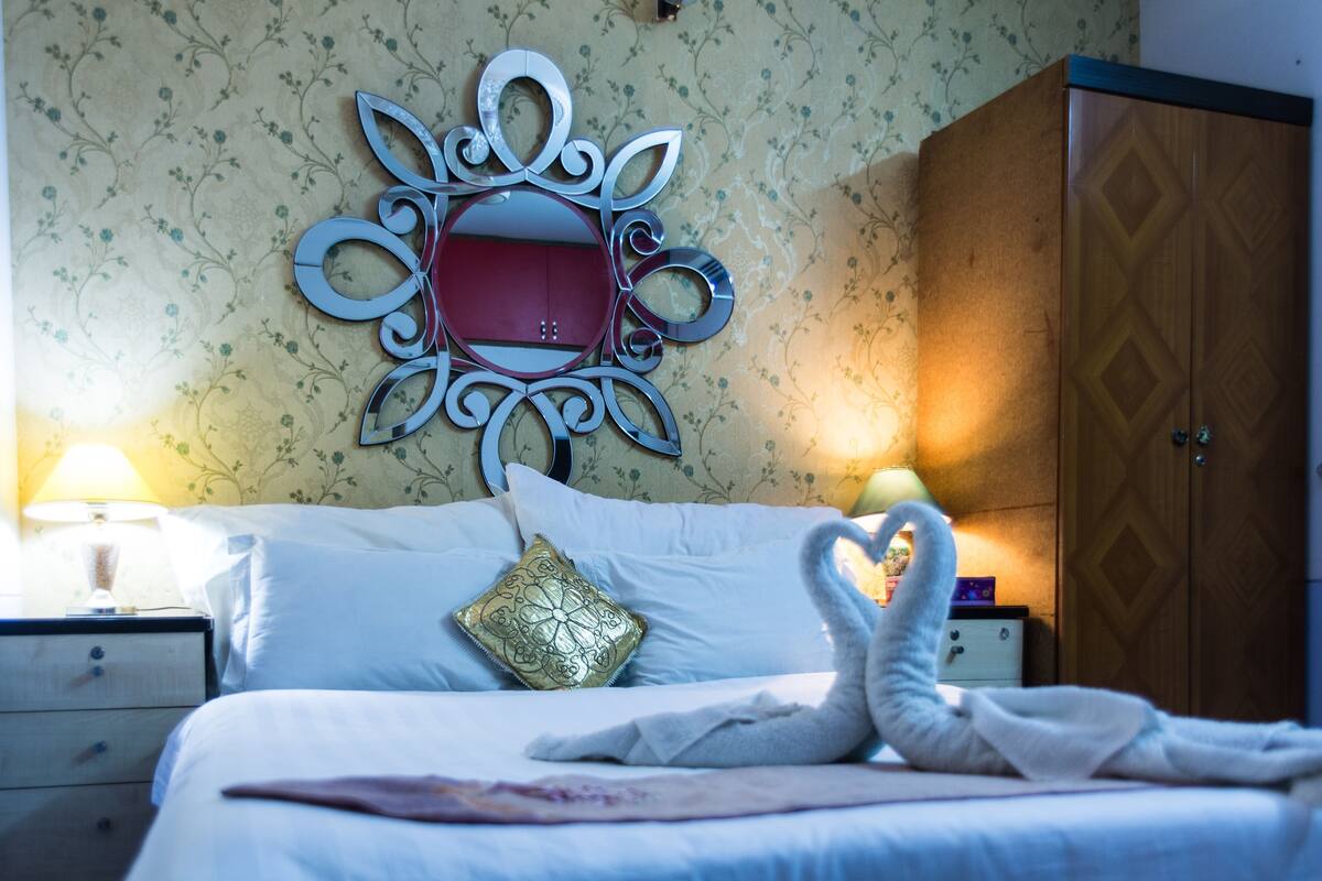A neatly made bed with a decorative mirror above it, a wooden wardrobe, and a nightstand with a lamp.