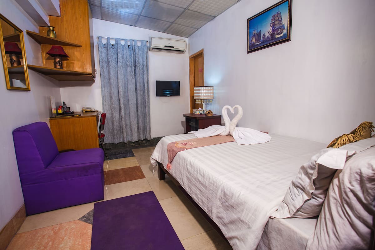 A hotel room with a purple sofa, a bed with white linens, a TV, a lamp, and a painting on the wall.